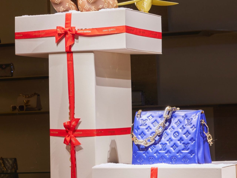 Oversized gift-box prop with a red ribbon beside a quilted handbag