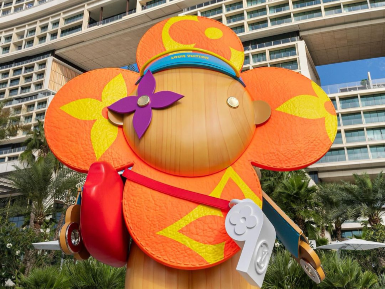 Large 3D-printed orange flower character sculpture with a red shoulder bag, installed outdoors beside a hotel